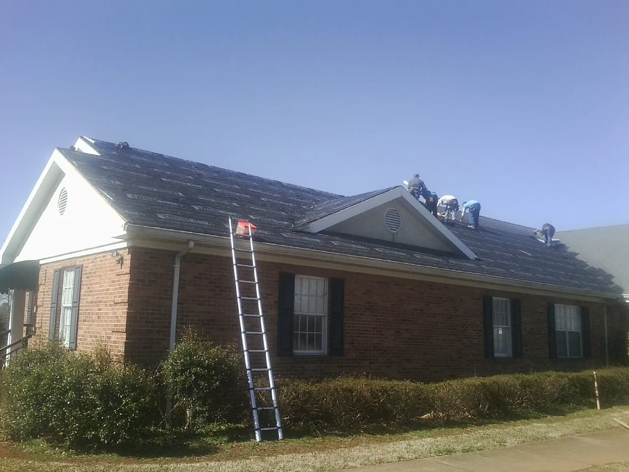 Crew tearing shingles off a brick ranch roof with a ladder set — Emergency Roof Repair work by our crew in Cadillac