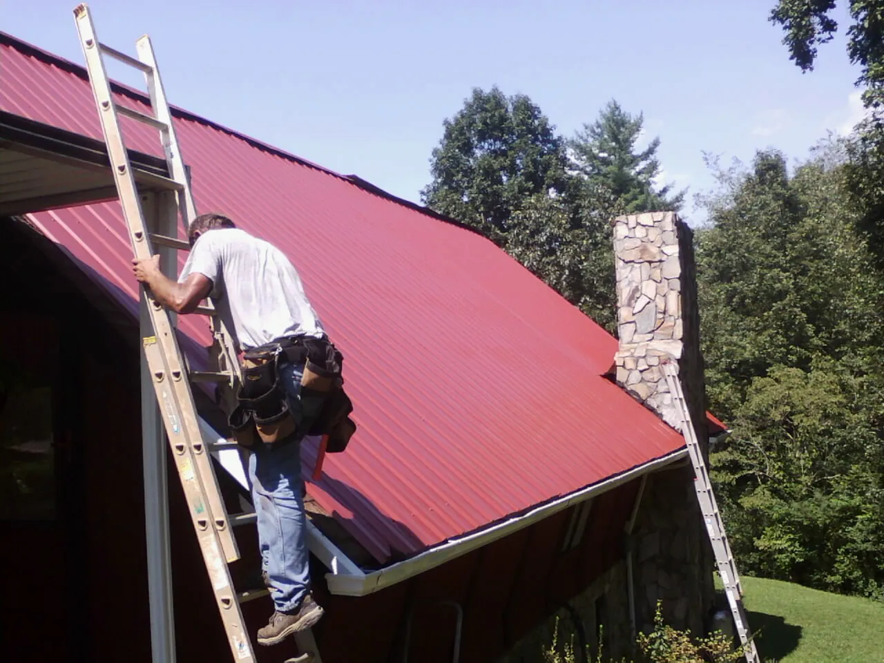 Roofer on a ladder beside a red metal roof and stone chimney — Soffit & Fascia Repair by Roofing Brothers in Cadillac